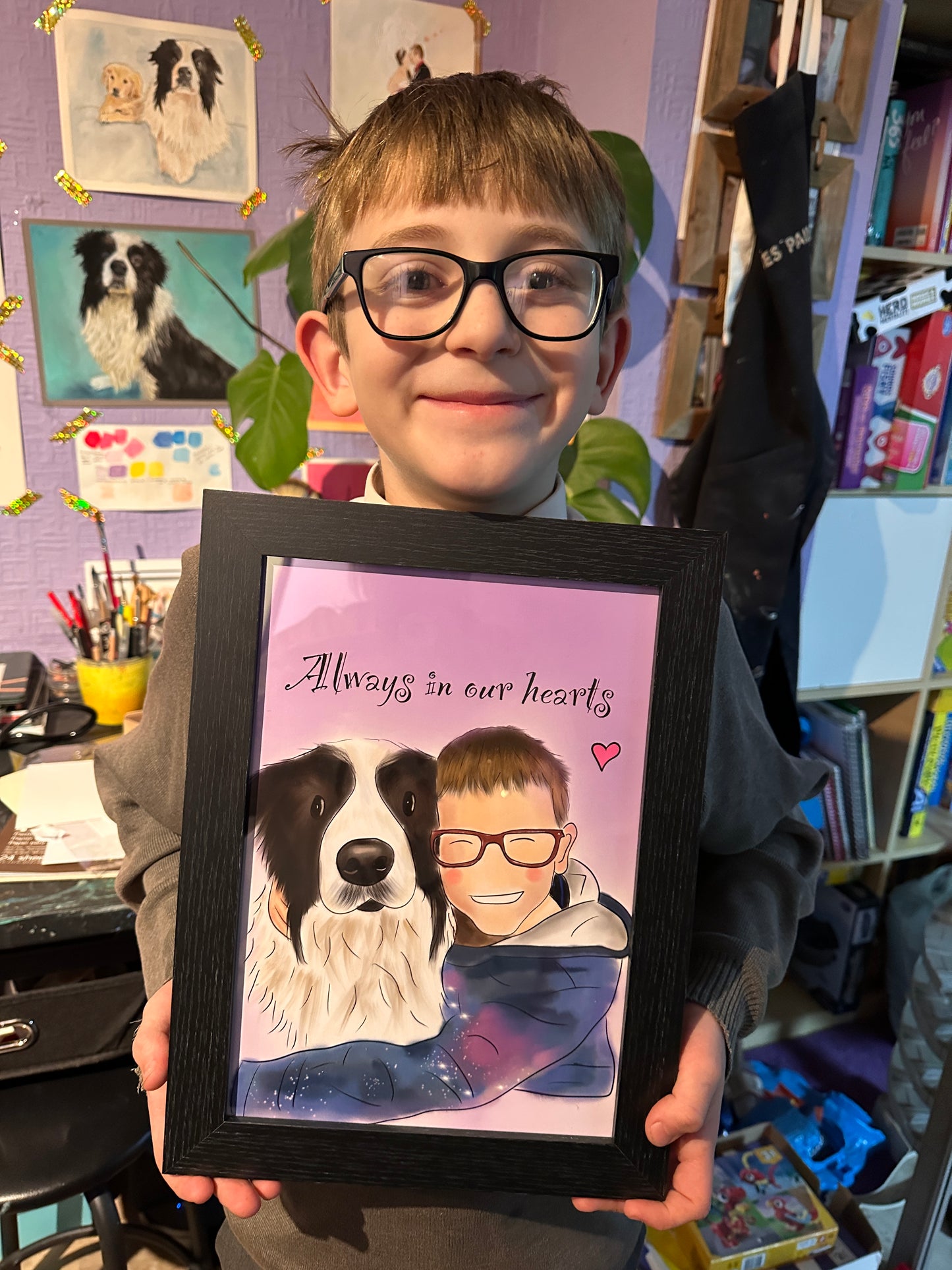 Child holding an a4 framed illustration of himself. The illustration is of a child and dog smiling together, hugging. It has a purple background, text that reads Always in our hearts with a pink heart in the right corner.