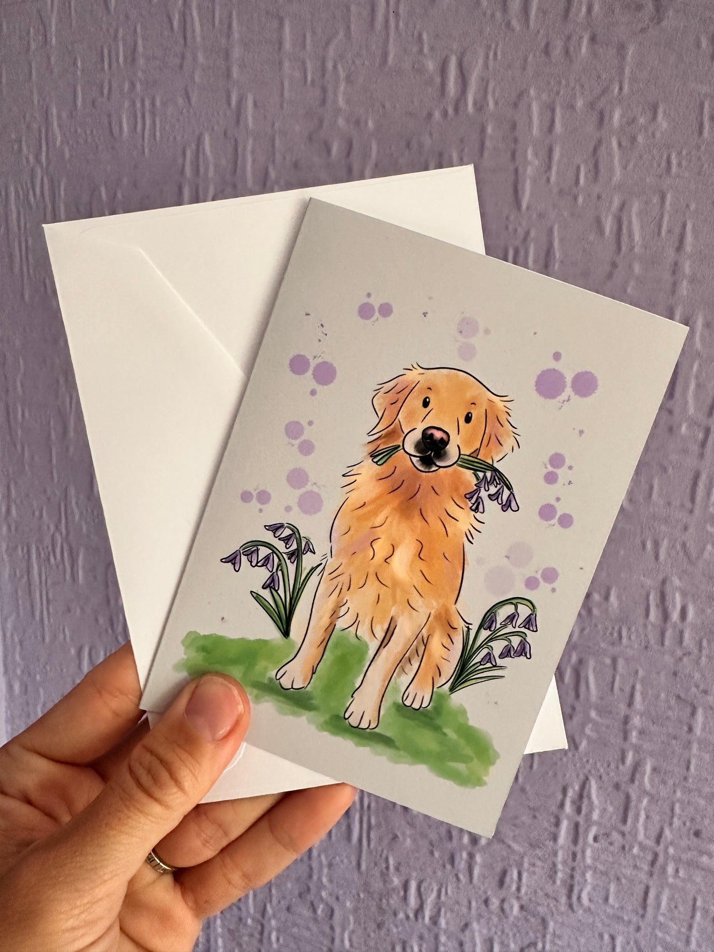 Greeting card and envelope being held up against a purple wall. Card has an illustration of a golden retriever sitting with bluebells in its mouth. It’s sitting on grass with bluebells either side of it and purple bubbles in the background.