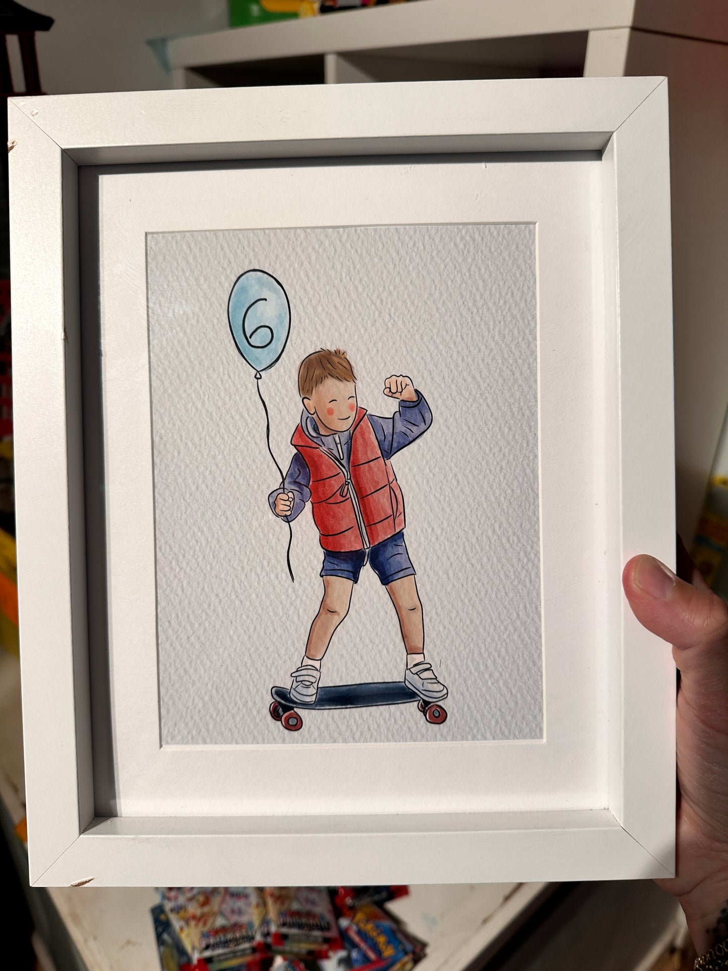 Framed picture of an illustration of a boy on a skateboard wearing shorts and a red jacket holding a blue balloon with a number 6 on it.