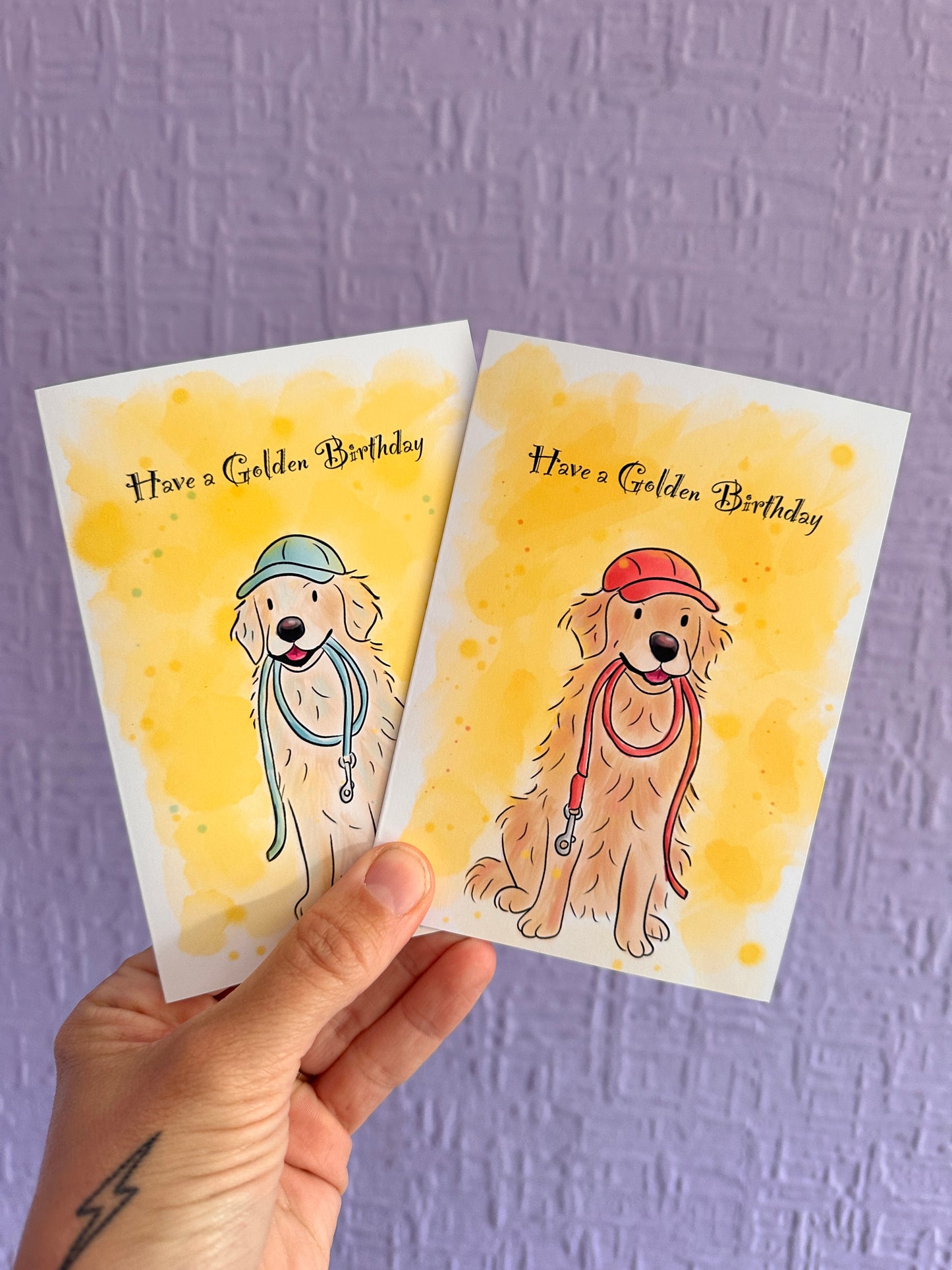 2 greeting cards being held up against a purple wall. The cards have a bright yellow water colour background and illustrations of golden retriever’s sitting looking happy. The left card the golden retriever is holding a blue lead and wearing a blue cap and the right card is a red lead and red cap. There is text saying Have a Golden Birthday.