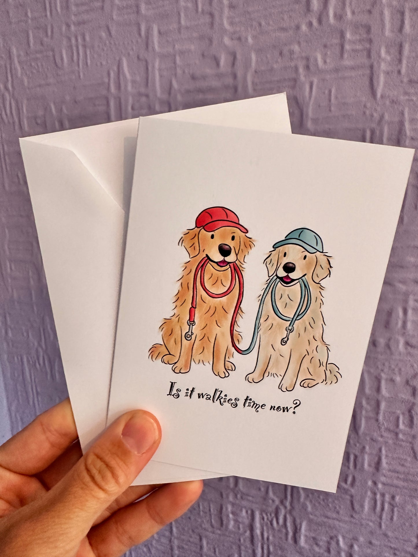 Greeting card and envelope being held up against a purple wall. Greeting card has illustrated golden retriever’s both sitting happy with leads in their mouths and wearing caps. The leads and caps are red and blue and there is text that reads Is it Walkies tie yet?