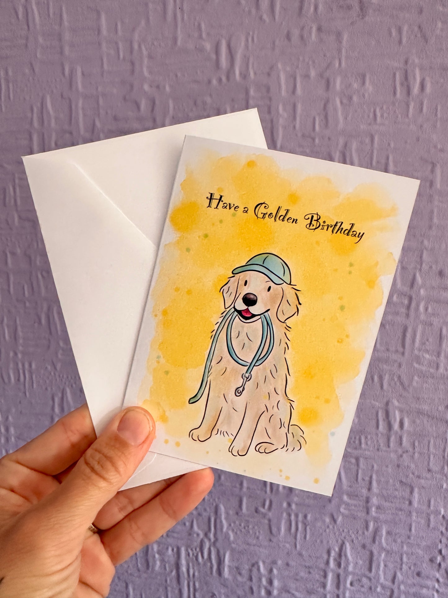 Greeting card and envelope of an illustrated golden retriever sitting with a blue lead in his mouth wearing a blue cap. Card had text saying Have a Golden Birthday, a Yellow background and is being held up by a purple wall.