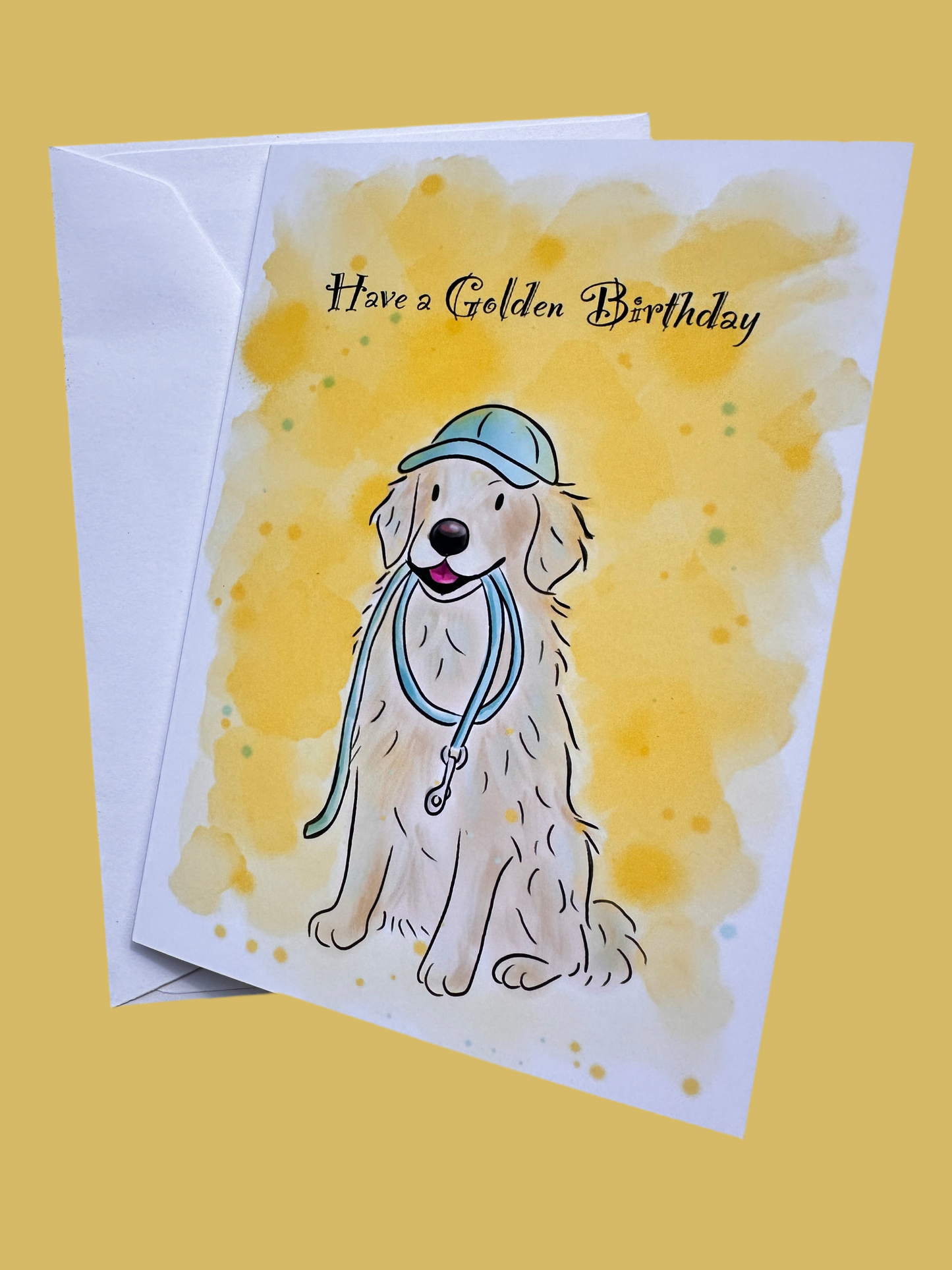 Greeting card and envelope with a yellow background. Card has an illustration of a golden retriever sat nicely wearing a blue cap and holding a lead in their mouth. The card has a bright yellow background and there is text that reads Have a Golden Birthday.