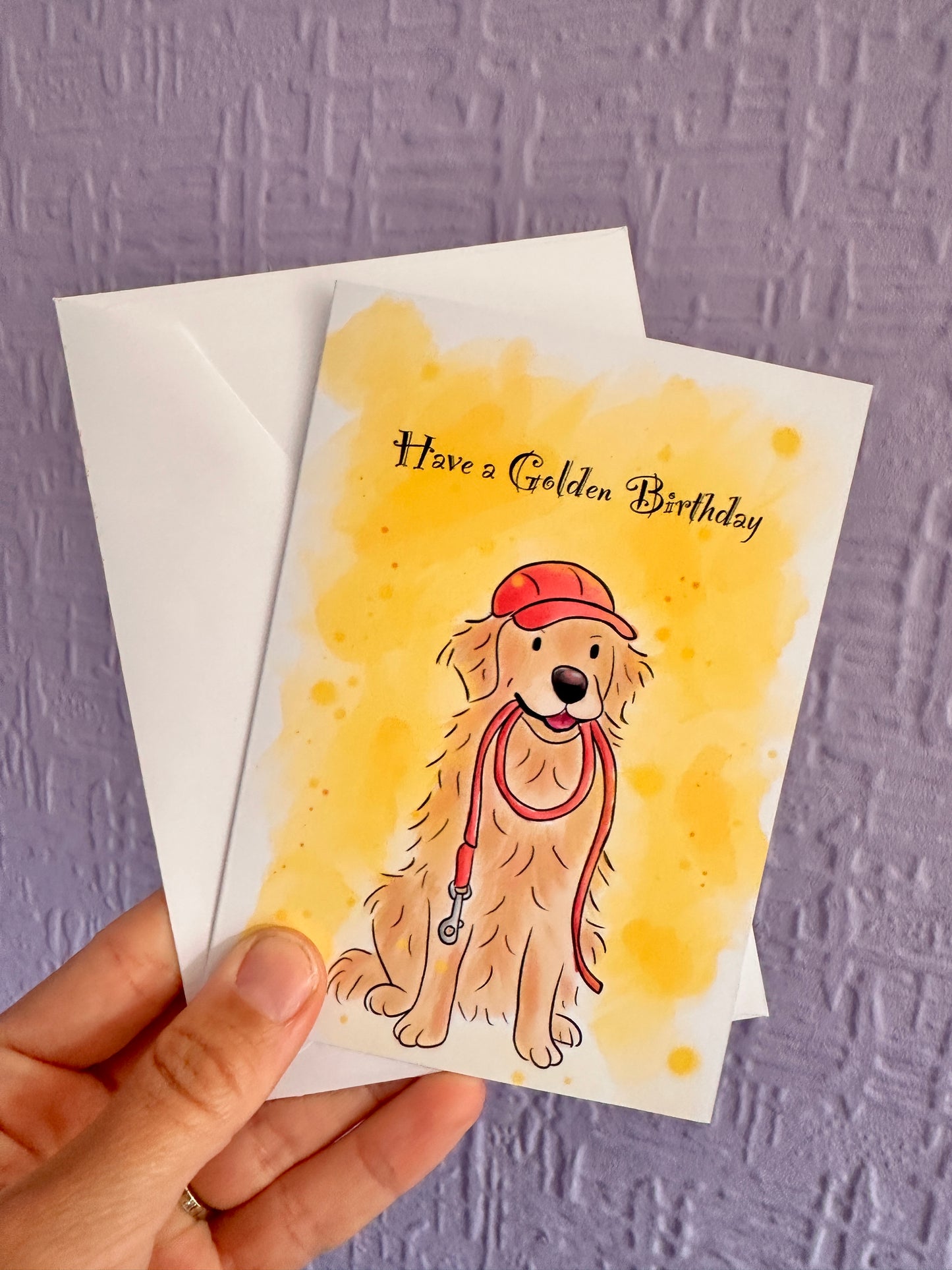 Greeting card held up by a purple wall with an envelope. The card has a golden retriever illustration where it is sitting happy holding a red lead in its mouth wearing a red cap. There is text that says Have a Golden Birthday. The card also has a bright yellow background.