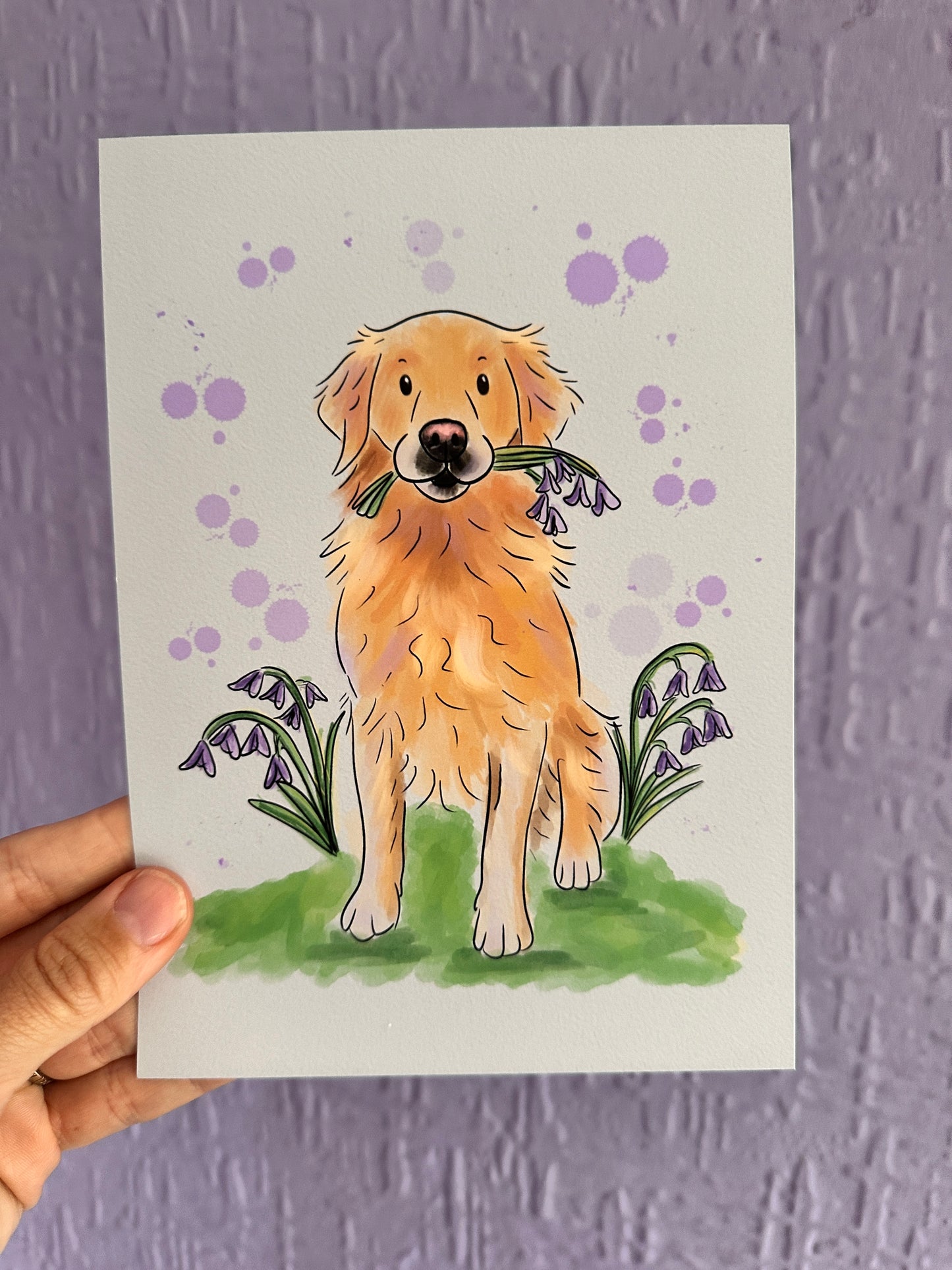 A golden retriever illustration art print being held up to a purple wall. The golden retriever is sitting in green grass and bluebells with bluebells in its mouth and purple bubbles in the background.