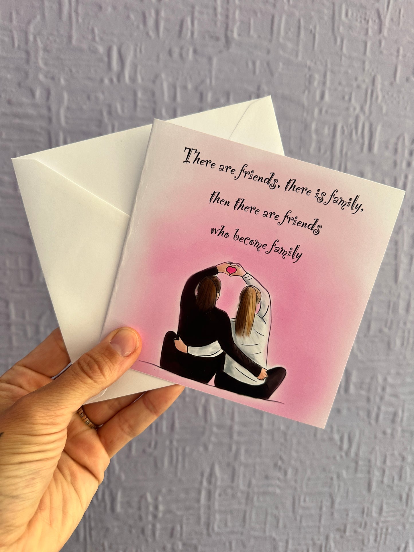 Image of a greeting card & envelope being held up against a purple wall. The card has an illustration of 2 girls cuddling sat next to each other with their arms up forming a heart with their hands. A pink background with text saying, there are friends, there is family, then there are friends who become family