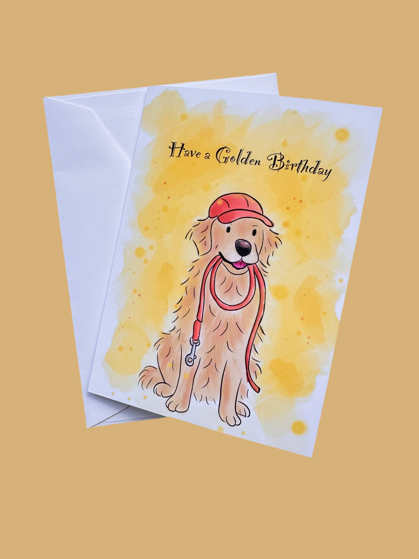 Golden background with Greeting card and envelope. Card has text saying Have a Golden Birthday, a bright yellow background and an illustration of a golden retriever sat with a red lead in its mouth wearing a red cap.