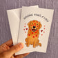 An image of a greeting card & envelope being held up against a purple wall. The card has an illustration of a sitting golden retriever which is covered in red lipstick kisses on its face & body and is surrounded by red hearts. The text at the top says SENDING KISSES & LOVE 