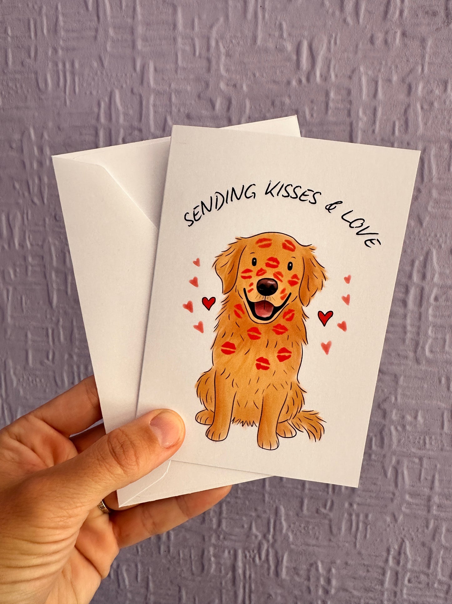 An image of a greeting card & envelope being held up against a purple wall. The card has an illustration of a sitting golden retriever which is covered in red lipstick kisses on its face & body and is surrounded by red hearts. The text at the top says SENDING KISSES & LOVE 