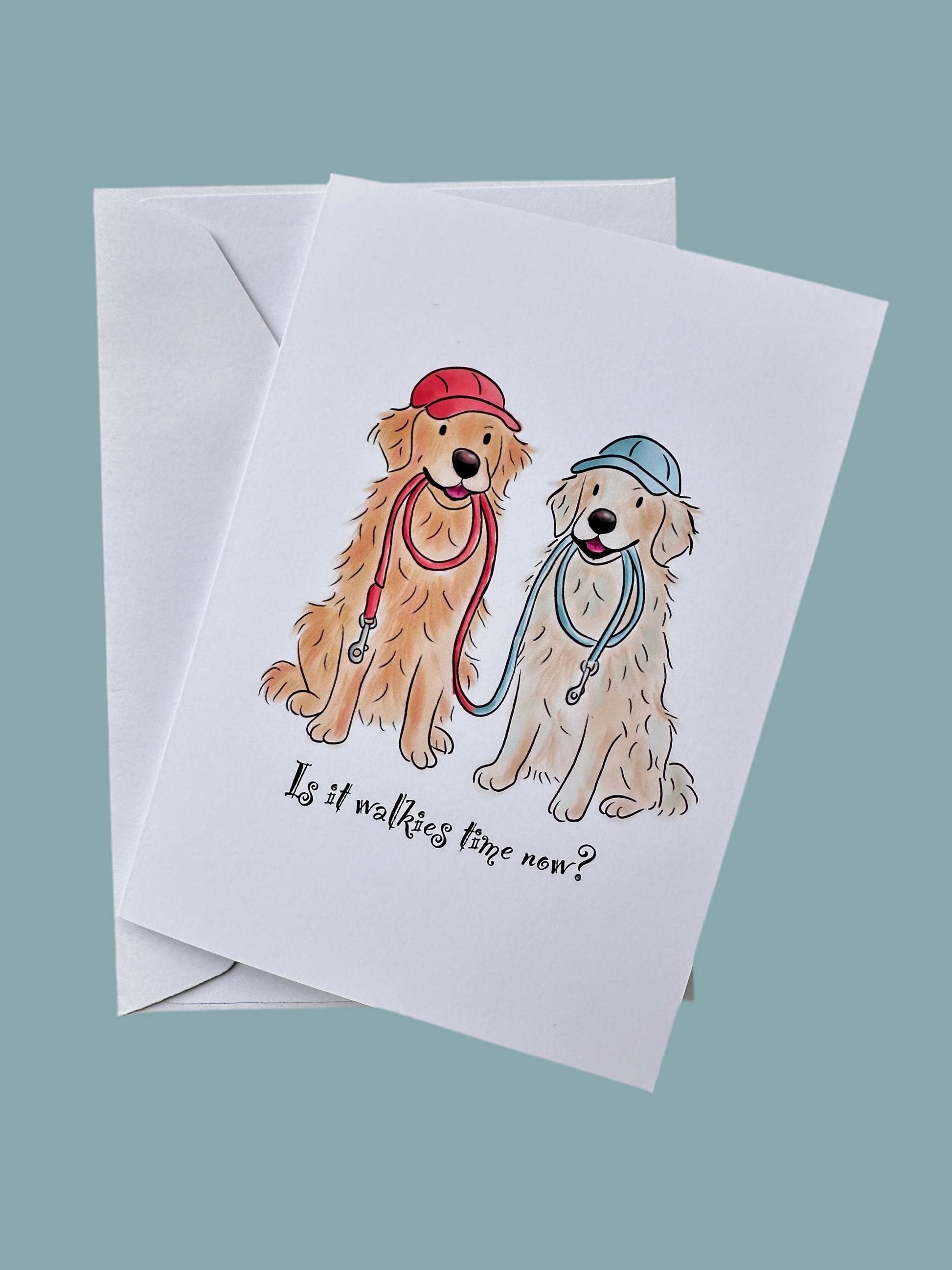 Greeting card with an illustration of 2 golden retrievers sitting with a red and blue lead in their mouths, wearing red and blue caps. The text says Is it walkies time yet and the background is blue. It also shows the envelope. 