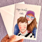 Illustration card and envelope being held up against a purple wall. Illustration of a mum with a big wide smile, bright sunglasses, pink cap and blonde hair hugging her kid from behind. Child is also smiling wearing a black and white football kit. Text says happy birthday Ollie. 
