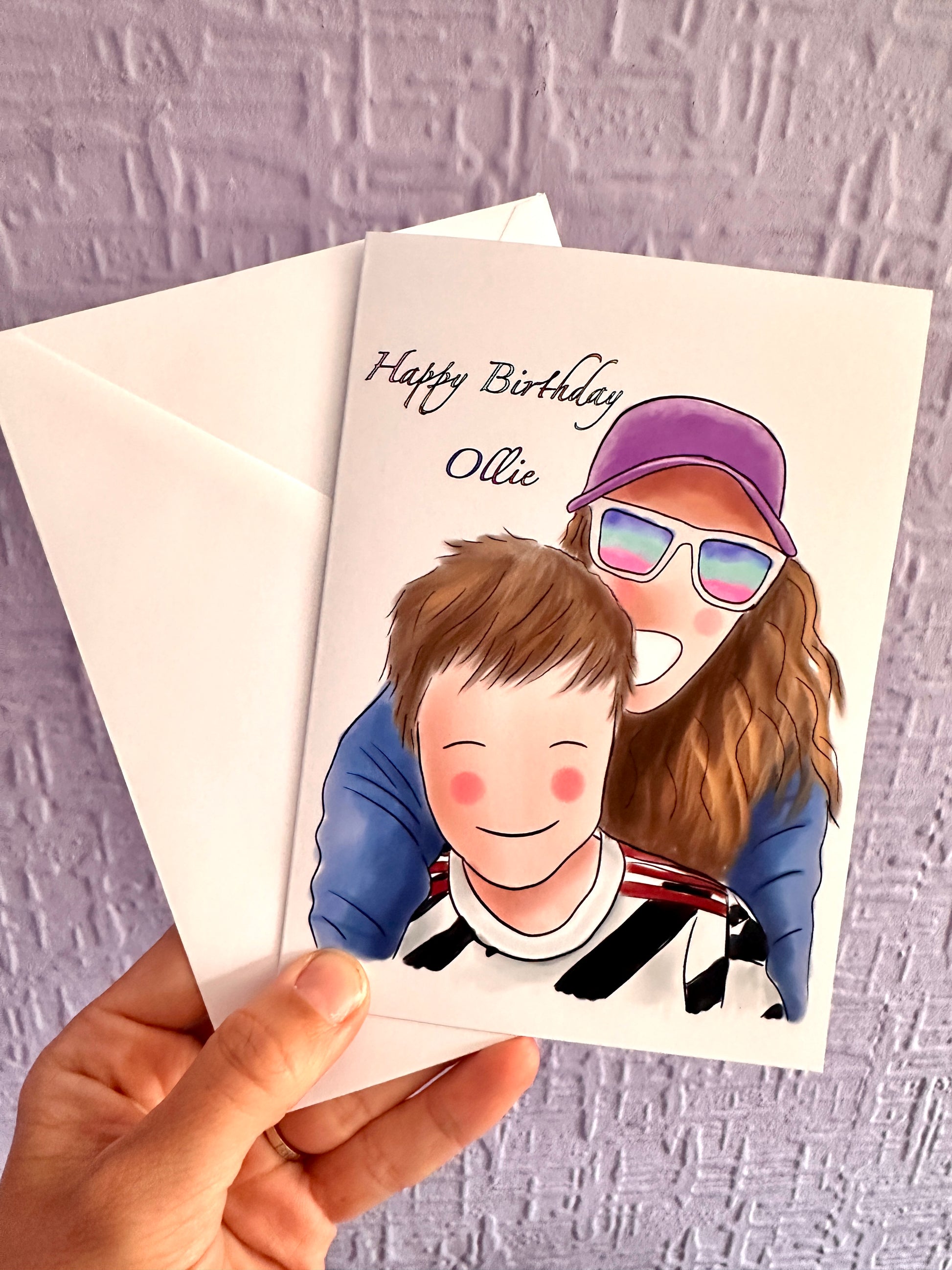 Illustration card and envelope being held up against a purple wall. Illustration of a mum with a big wide smile, bright sunglasses, pink cap and blonde hair hugging her kid from behind. Child is also smiling wearing a black and white football kit. Text says happy birthday Ollie. 