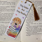 Bookmark laying on the middle of an open book with an illustration of a golden retriever leaning on a stack of rainbow books. There is text on the bookmark saying Stay golden And Keep Reading, there are rainbow stars and a golden tassel. 