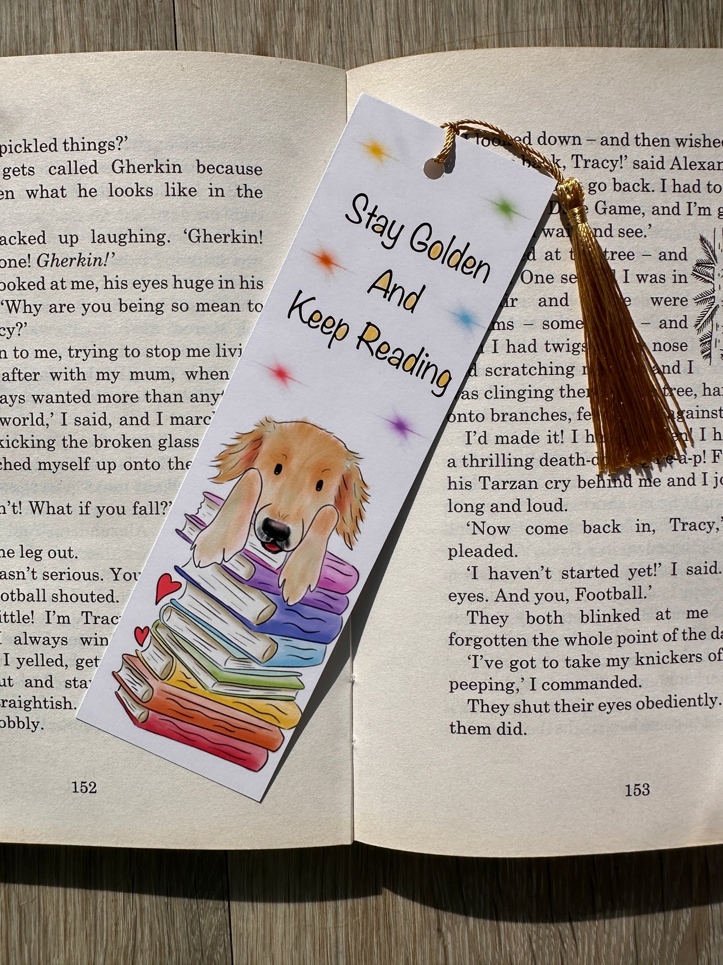 Bookmark laying on the middle of an open book with an illustration of a golden retriever leaning on a stack of rainbow books. There is text on the bookmark saying Stay golden And Keep Reading, there are rainbow stars and a golden tassel. 