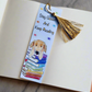 Bookmark is resting on a plain open book. The bookmark has a golden tassel attached, text saying Stay Golden And Keep Reading, rainbow stars on the background and an illustration of a golden retriever on top of a stack of rainbow books. 