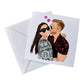 Square illustration card plus envelope of a couple hugging. Woman has long brown hair, sunglasses, big smile and black/white top. Man has blonde hair, black red top and is kissing her cheek. There are 2 pink love hearts in the background. 