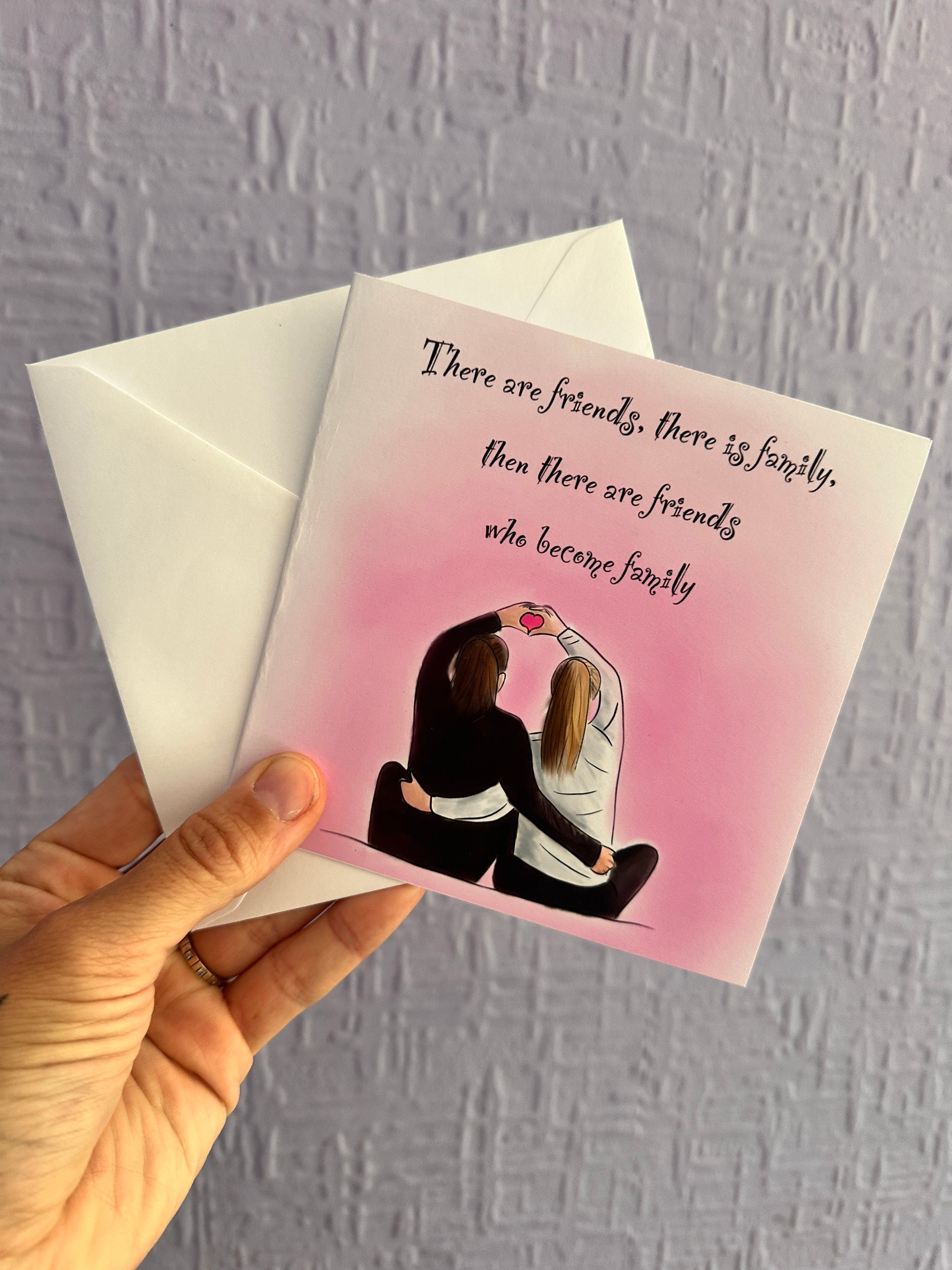 Image of a greeting card & envelope being held up against a purple wall. The card has an illustration of 2 girls cuddling sat next to each other with their arms up forming a heart with their hands. A pink background with text saying, there are friends, there is family, then there are friends who become family 