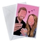Illustration card plus envelope of a couple looking happy together holding shots of drinks. Man has a beard and a black shirt, woman has long blond hair and denim jacket. There’s 3 pink love hearts and a pink background and think white border. 